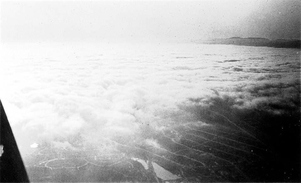 An aerial view looking past an aircraft strut at a thick blanket of white clouds, with a patch of coastal land visible below.