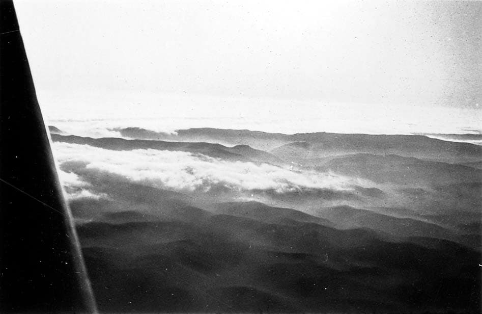 An aerial view looking past an aircraft strut at a thick blanket of clouds rolling over hills and mountains.
