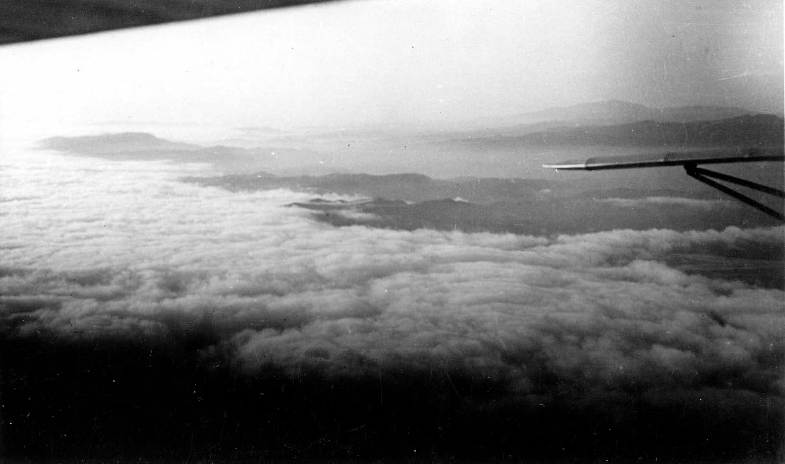 An aerial view looking out over a vast sea of clouds stretching toward a mountainous horizon, framed by an aircraft strut.