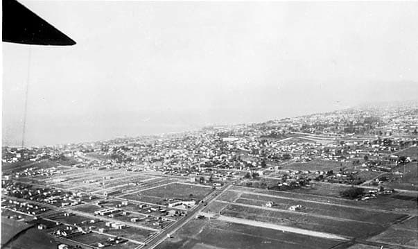 An aerial view looking out from under an aircraft wing toward a sprawling coastal city grid bordering the Pacific ocean.