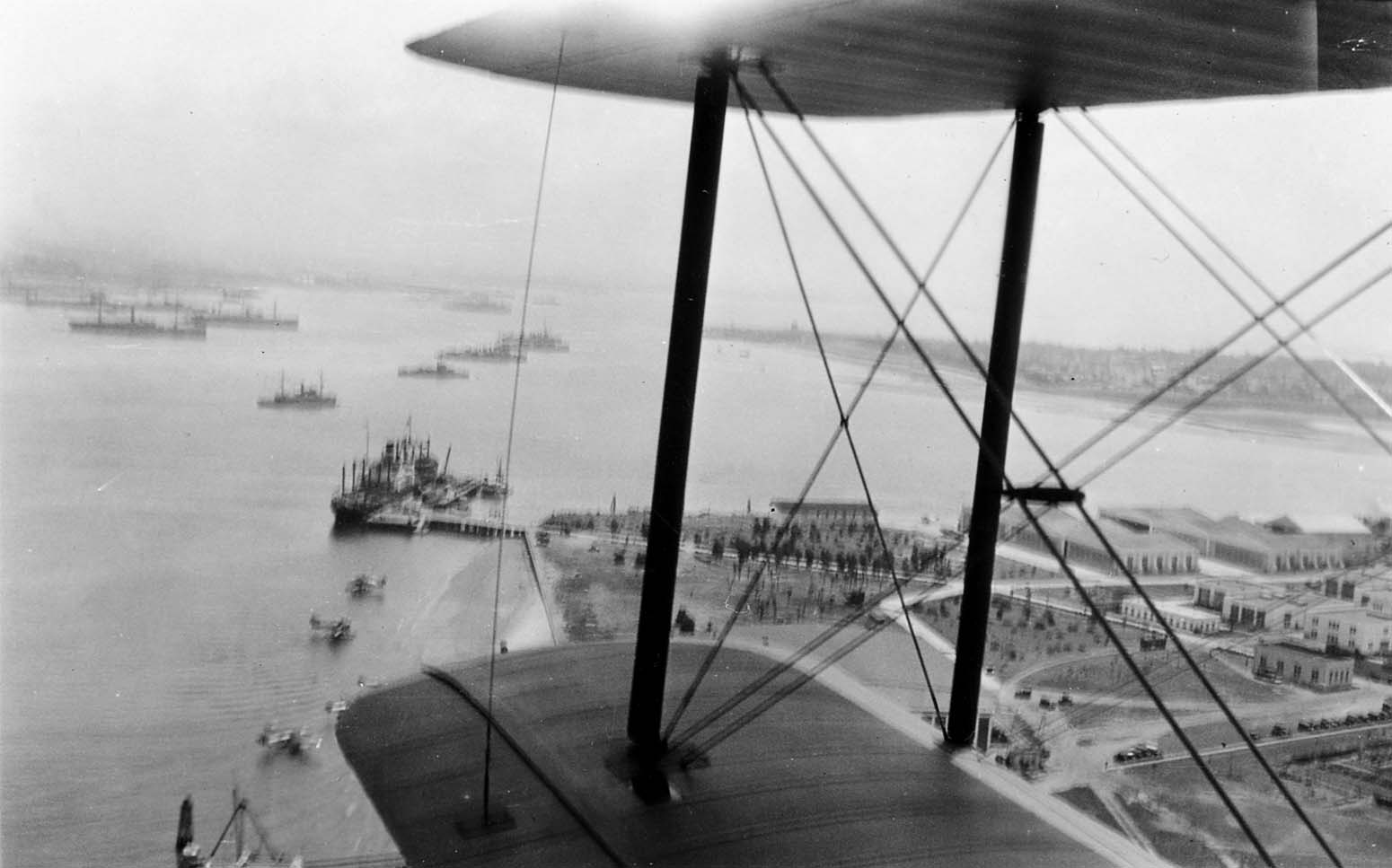 Looking down past the lower wing and struts of a biplane at a coastal harbor filled with anchored ships and a pier.