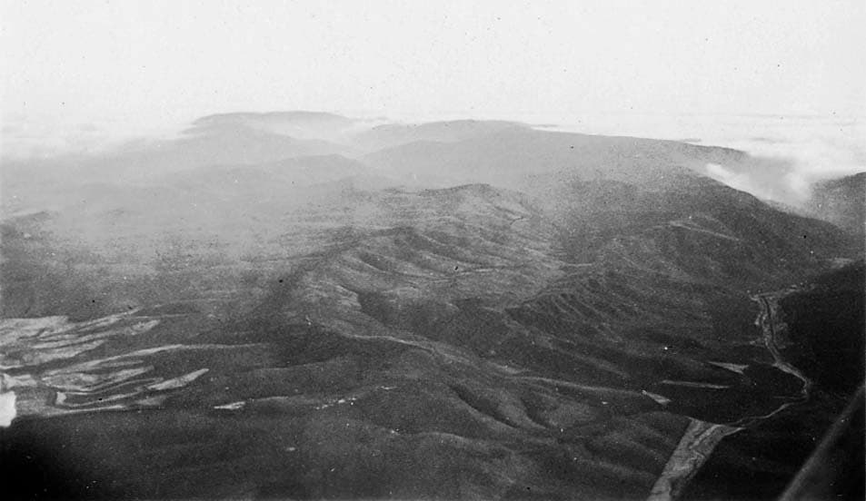 An aerial photograph showing a vast expanse of rolling hills and valleys beneath a layer of high clouds.