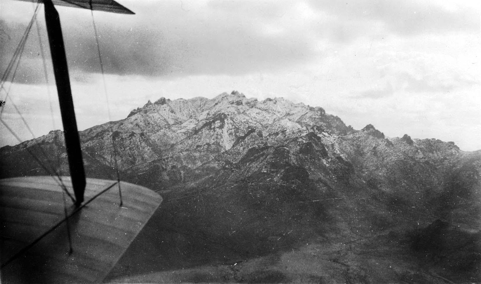 An aerial view looking past the trailing edge of a biplane wing toward a massive, rugged mountain peak dusted with snow.