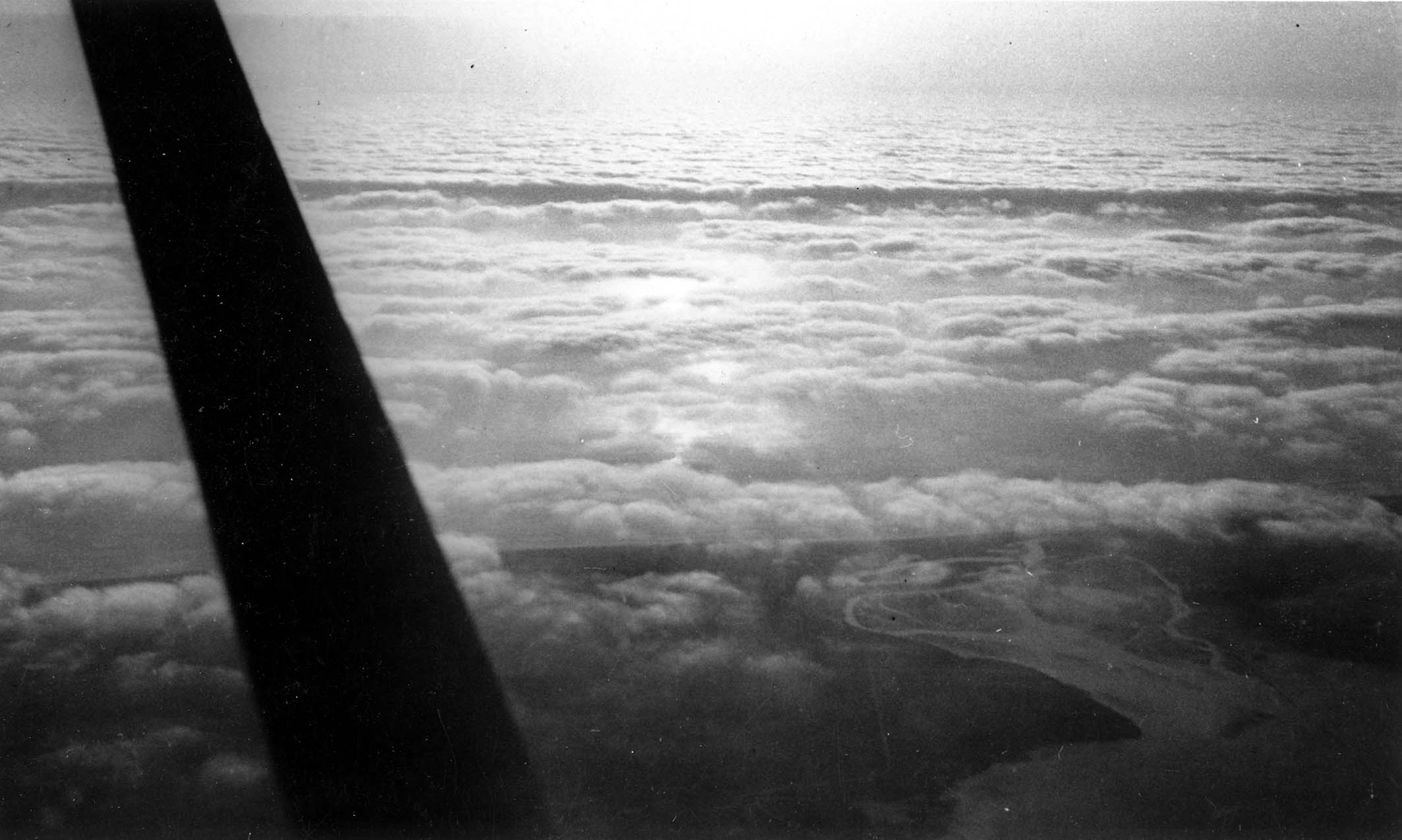 An aerial view looking past a wing strut down at a blanket of clouds, with a winding river delta visible through a break.