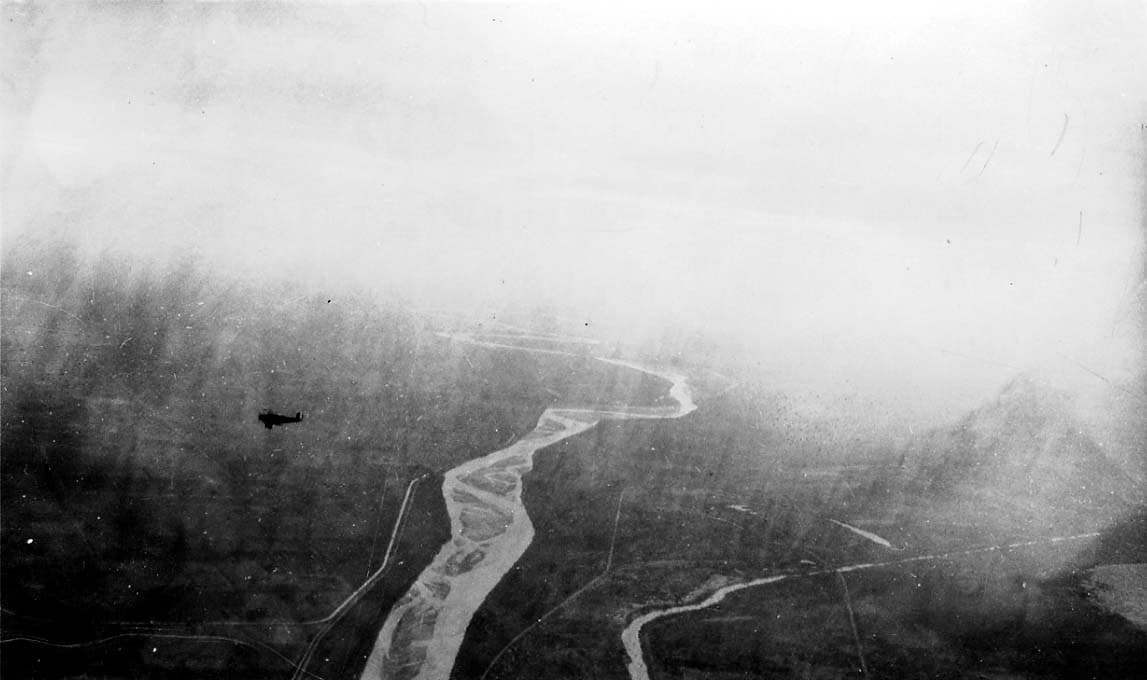An aerial view of a winding river valley shrouded in mist, with a small silhouette of another airplane flying in the distance.