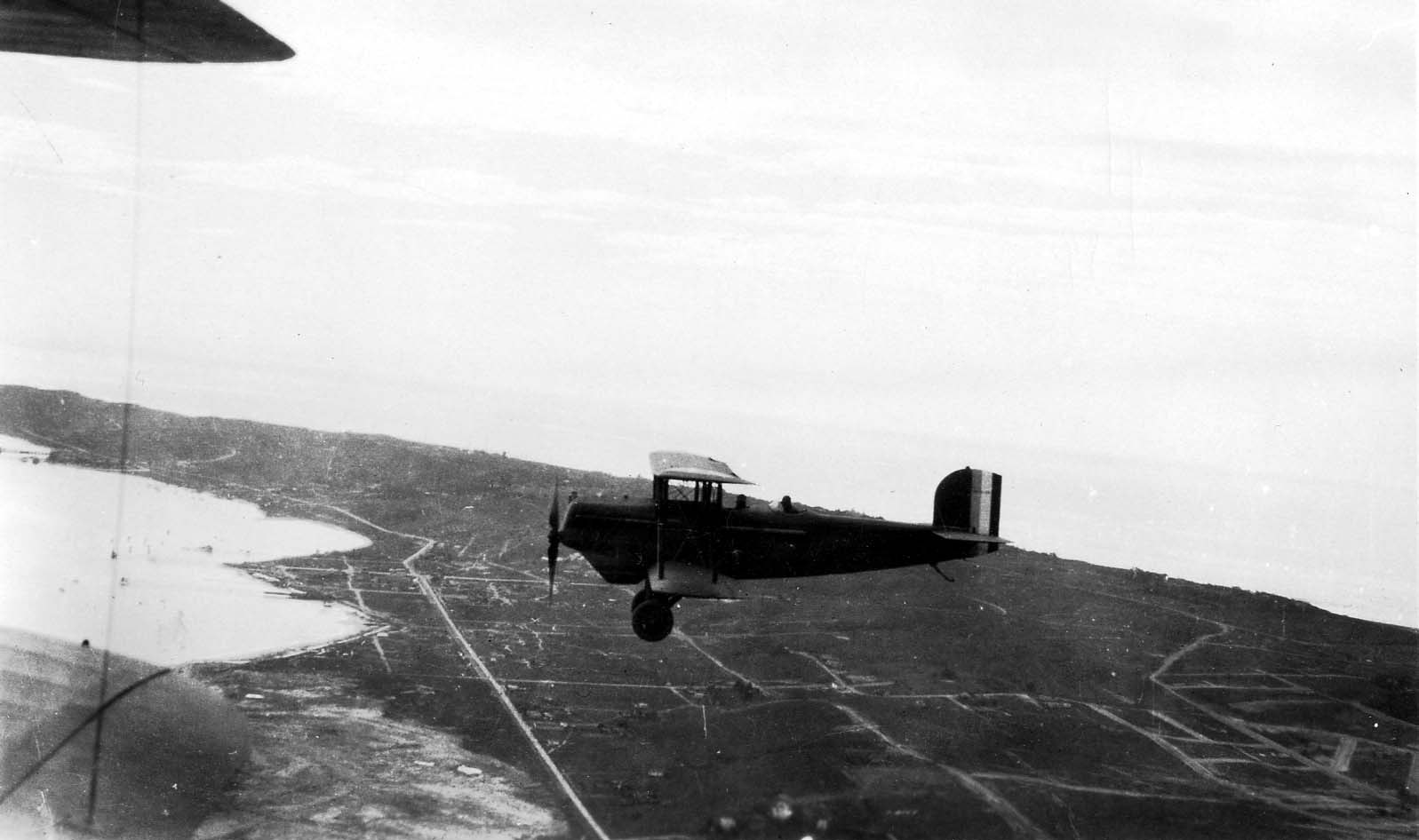 An air-to-air photograph of a biplane flying over a coastline with a bay and peninsula in the background.