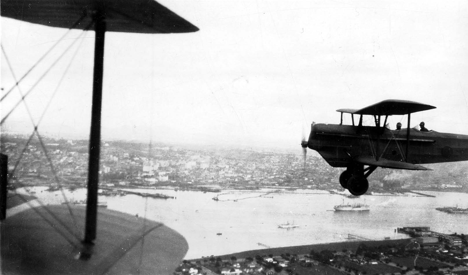 An incredible air-to-air photograph of a second biplane flying in formation over a harbor, taken from the cockpit of the lead aircraft.