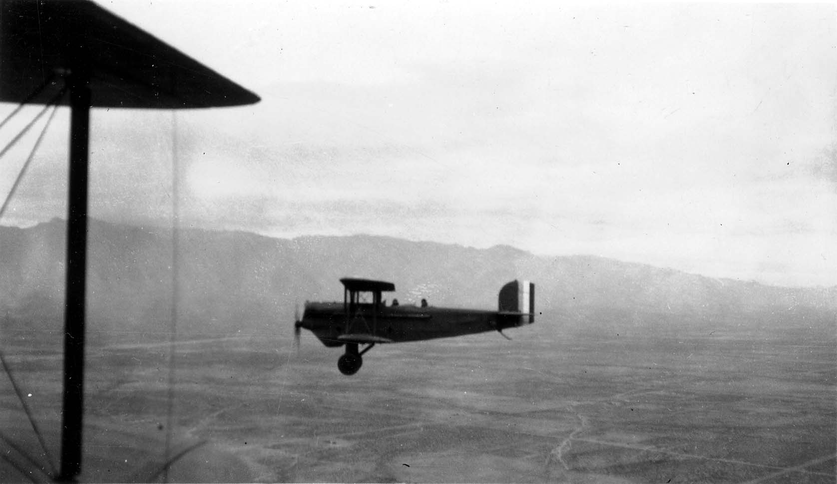An air-to-air photograph of a biplane in mid-flight against a mountainous background, taken past the wing struts of the camera plane.