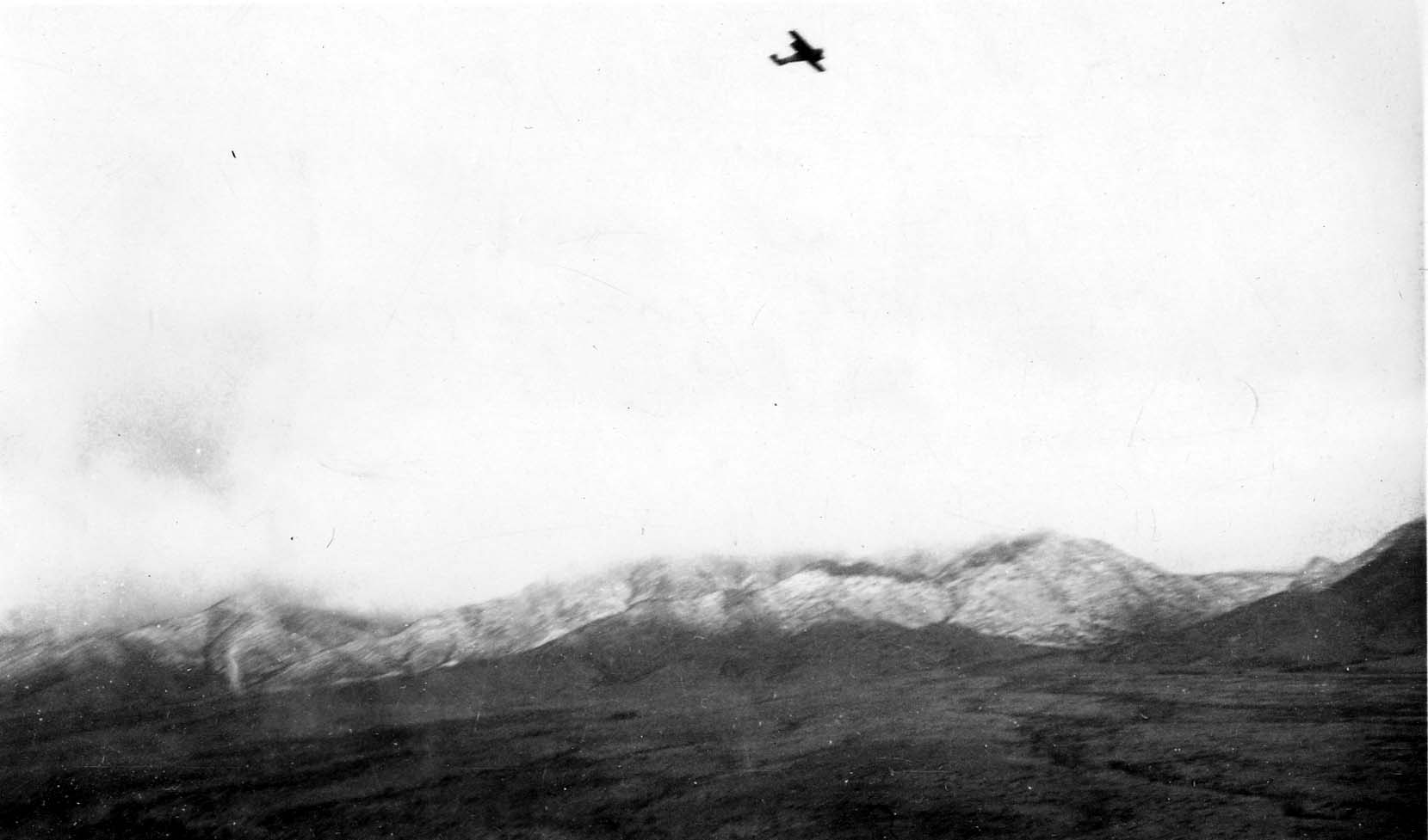 A distant view of an aircraft flying high above a rugged mountain range covered in fresh snow, captured from the air.