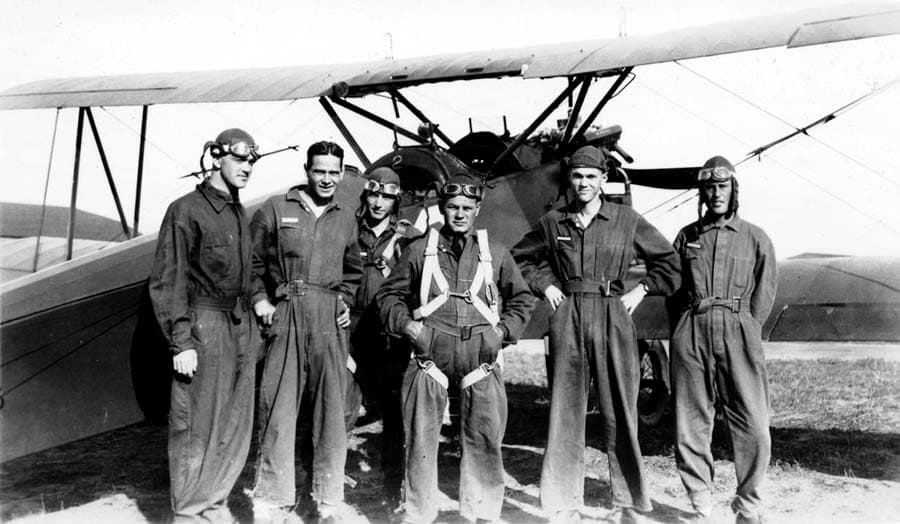 A group of five Army Air Corps flight cadets and an instructor standing in front of a biplane trainer on a dirt airfield.