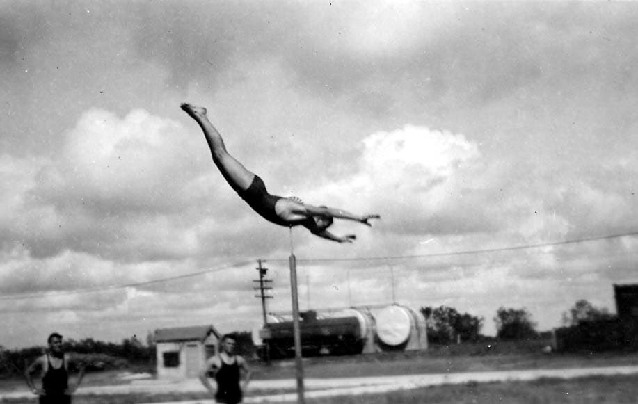 An incredible action photograph of a man executing a perfect swan dive off a high pole structure, with two men watching from the ground near a small building.