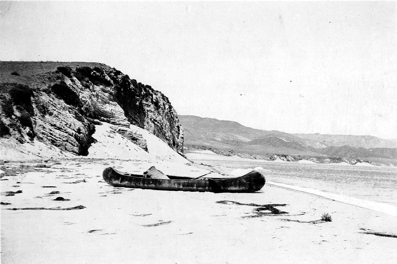 Donald A. Hall Sr.'s wooden canoe resting on a sandy beach at the base of towering stratified ocean bluffs, with calm sea water in the background.