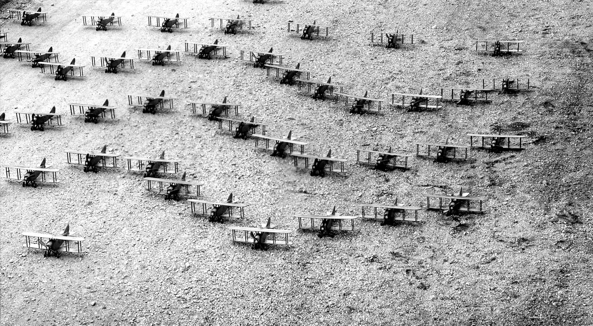 Dozens of miniature biplane models meticulously arranged in a V-formation flight pattern on the dirt ground.