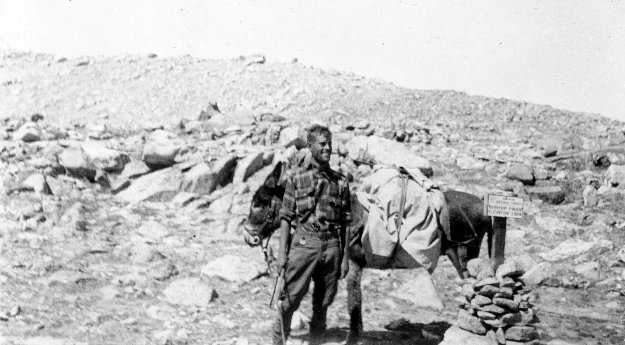 Donald A. Hall Sr. smiling next to a loaded pack mule at a wooden trail sign reading Bishop Pass.
