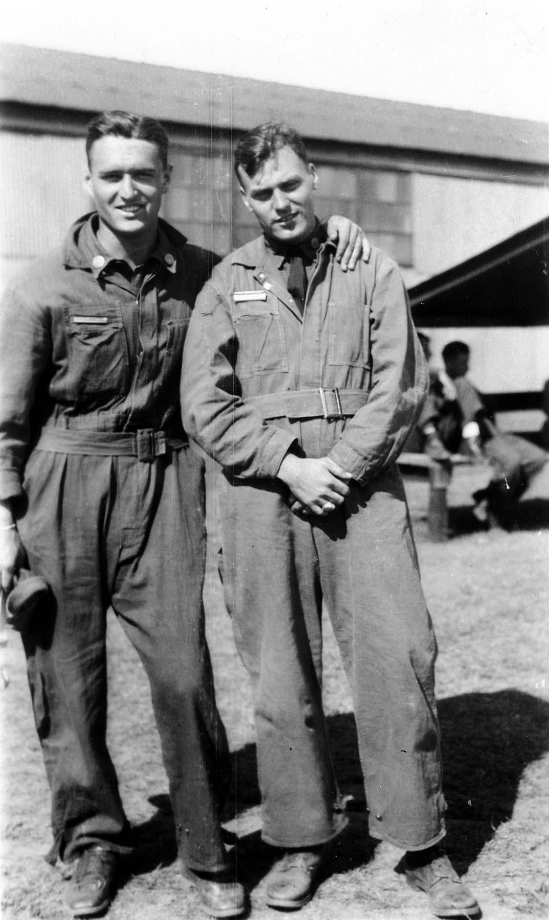 Two Army Air Corps flight cadets in military training coveralls smiling and posing together on the grassy airfield.