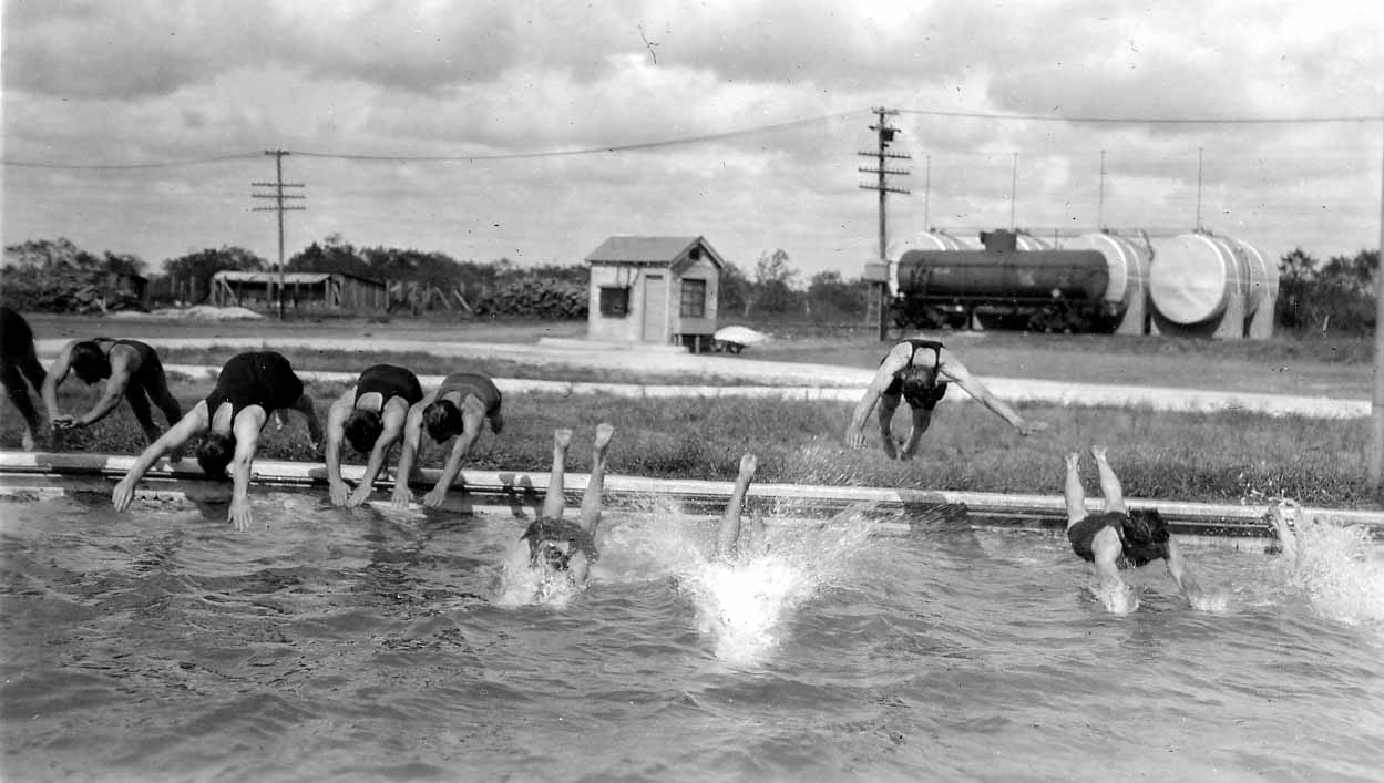 A group of six flight cadets diving simultaneously off the edge of the Brooks Field swimming pool, with fuel tanker cars parked on tracks behind them.