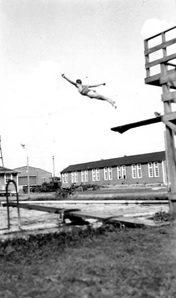 An action shot of a diver leaping high off a wooden springboard, arms outstretched, with the Brooks Field barracks in the background.