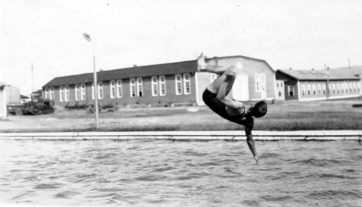 A diver arched backward in mid-air, about to enter the swimming pool, with a tractor and the long Brooks Field barracks visible in the background.
