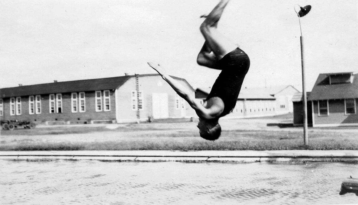 A close-up action photograph of a man frozen upside down in mid-air as he executes a diving flip, with Brooks Field military barracks in the background.