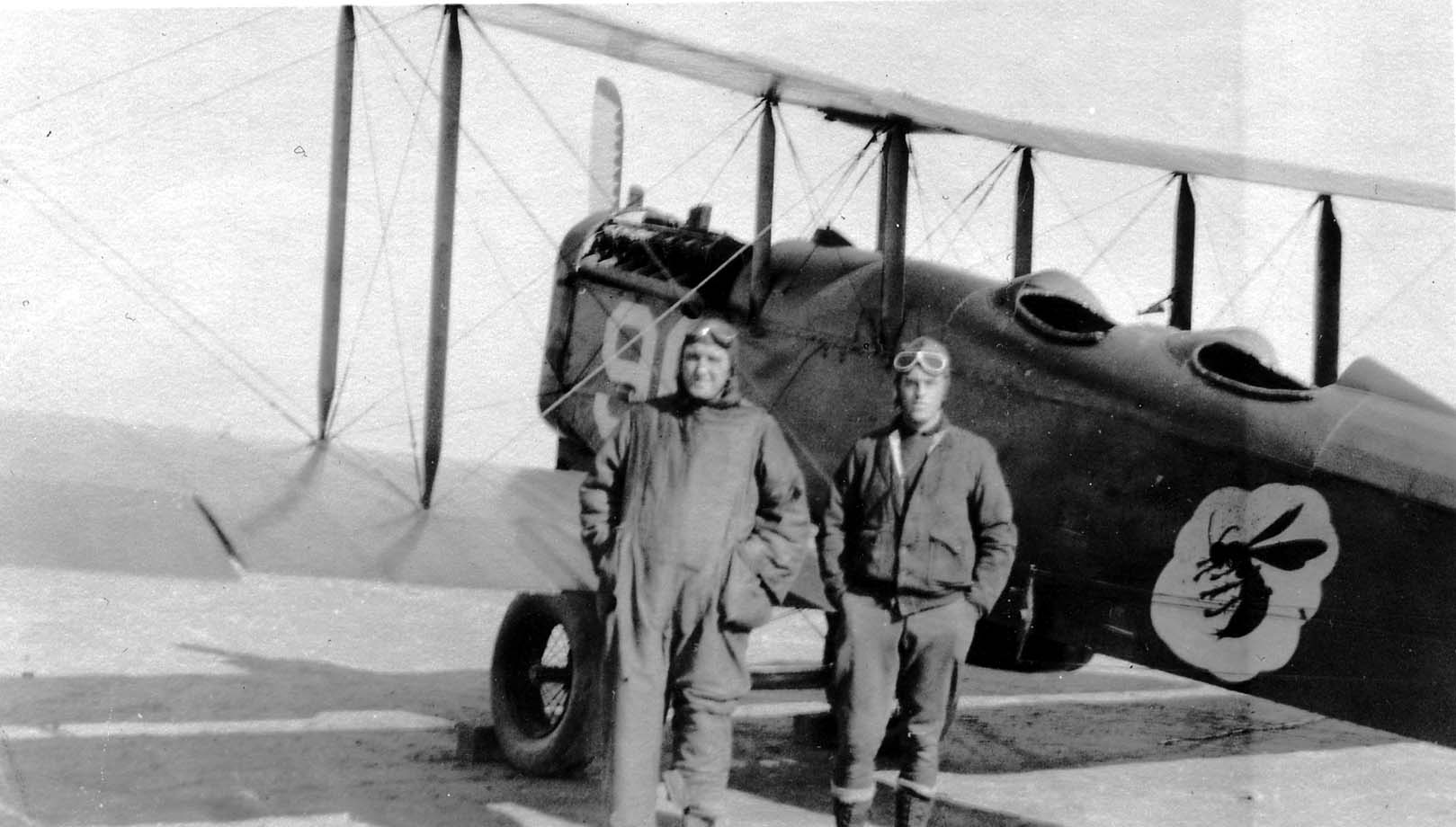 Two men in flight gear standing in front of an Army trainer biplane featuring a large insect emblem at Brooks Field.