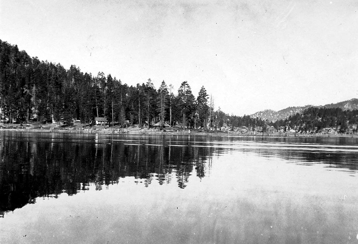 A calm mountain lake reflecting a dense forest and rustic lakeside cabins along the opposite tree-lined shore.