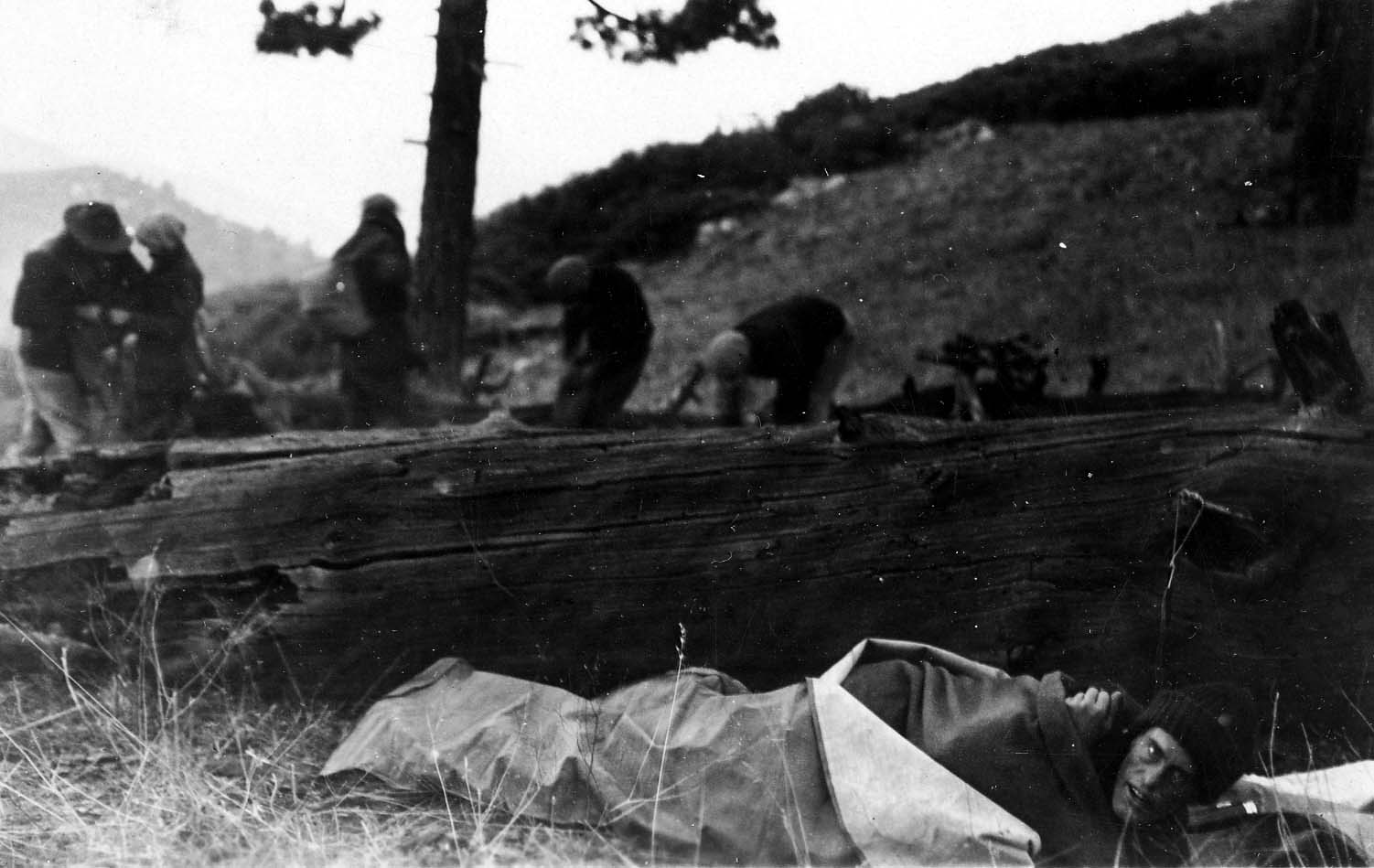 Donald A. Hall Sr. wrapped in a sleeping bag on the ground next to a massive fallen log, with other hikers in the background.