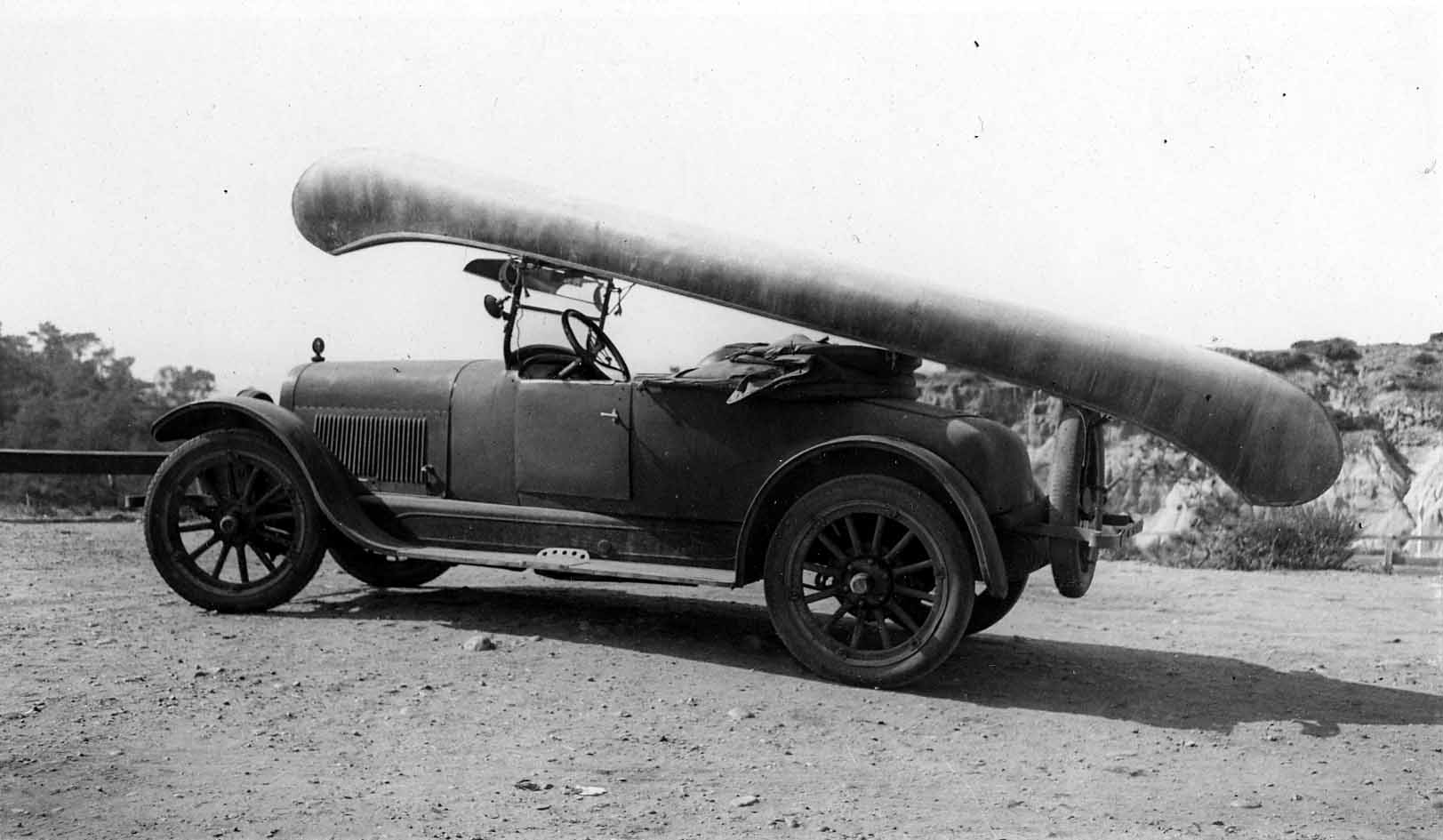 A large canoe tied upside down across the top of a vintage, open-cab automobile parked on a dirt road near a coastal cliff.