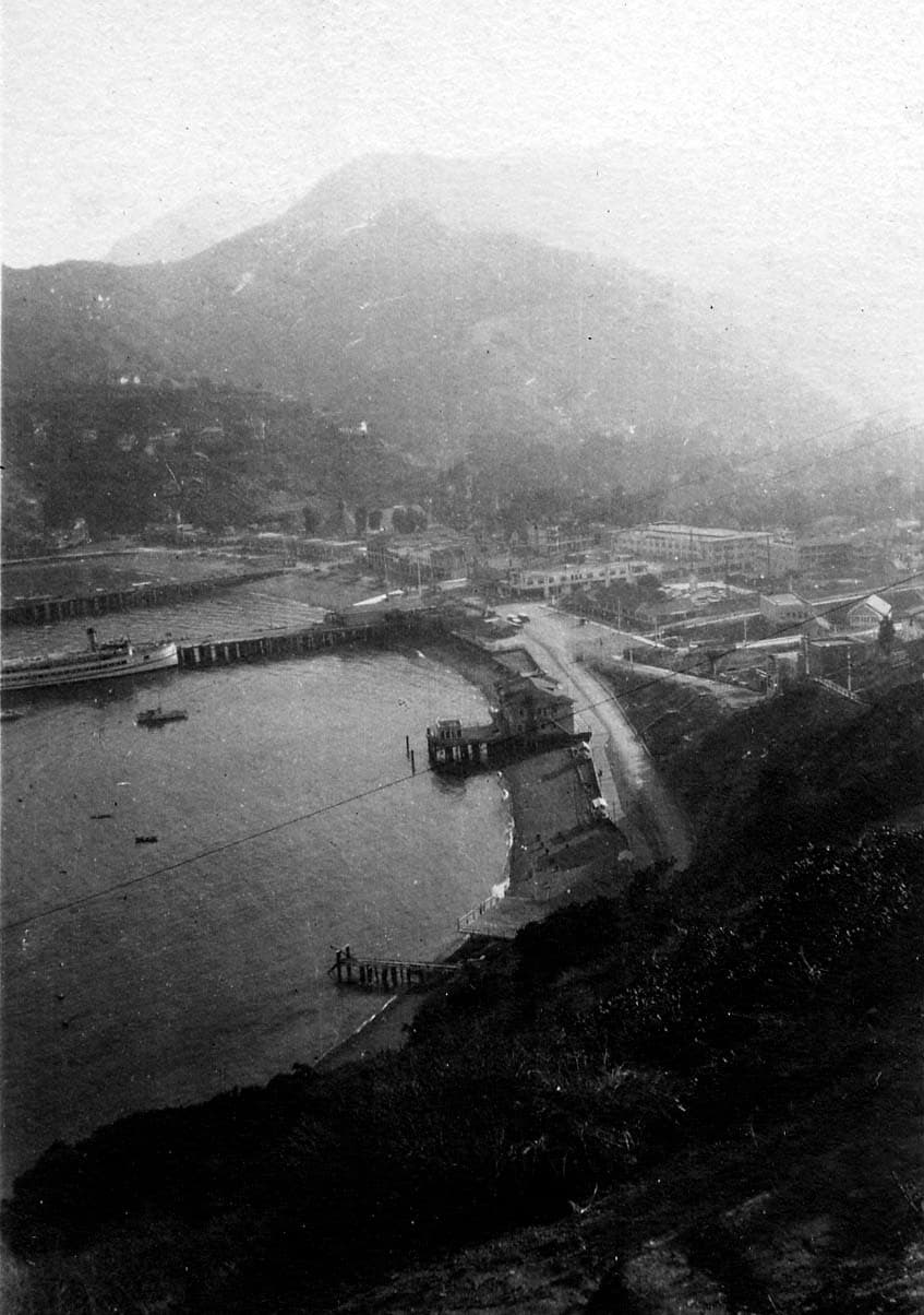 A high-altitude, hazy aerial view looking down a steep, scrub-covered hillside toward a coastal resort and a long pier jutting into a calm bay.