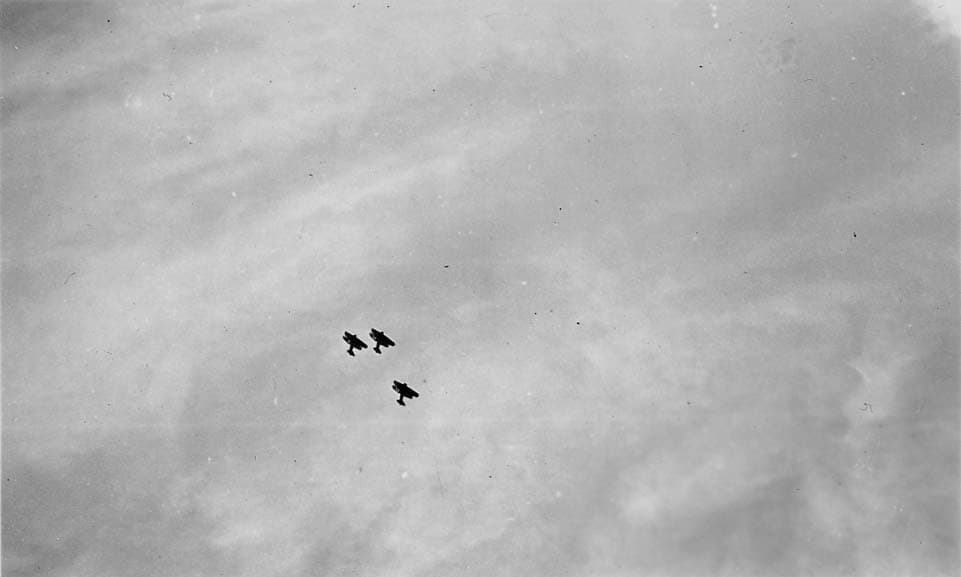 Three vintage biplanes flying in a tight V-formation high against a hazy, cloudy sky.