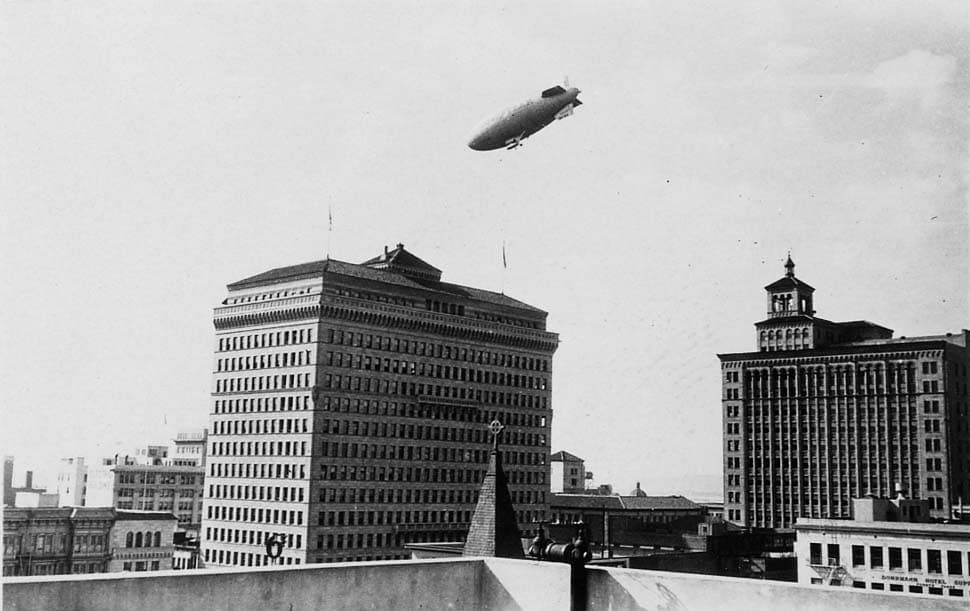 A vintage airship (dirigible) flying high above a dense city skyline featuring ornate, multi-story commercial buildings.