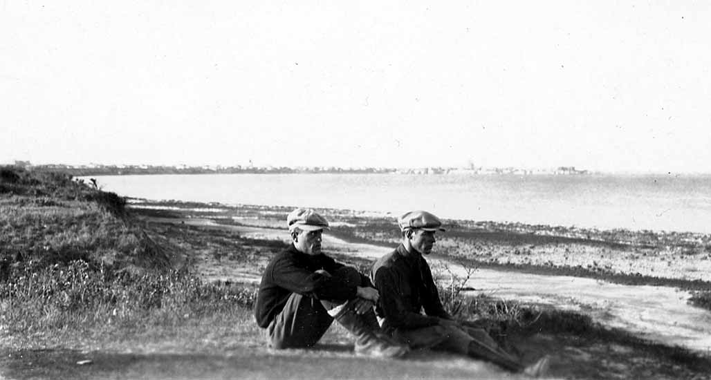 Two men sitting on a grassy coastal bluff looking out over the bay toward a distant cityscape.