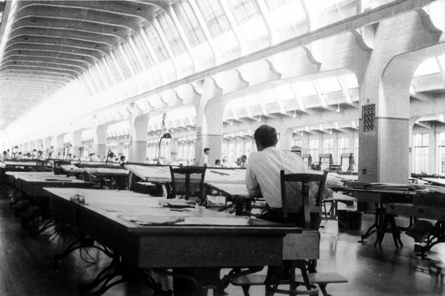 A massive, brightly lit industrial drafting room featuring rows of large drafting tables, massive arched concrete pillars, and extensive skylights.