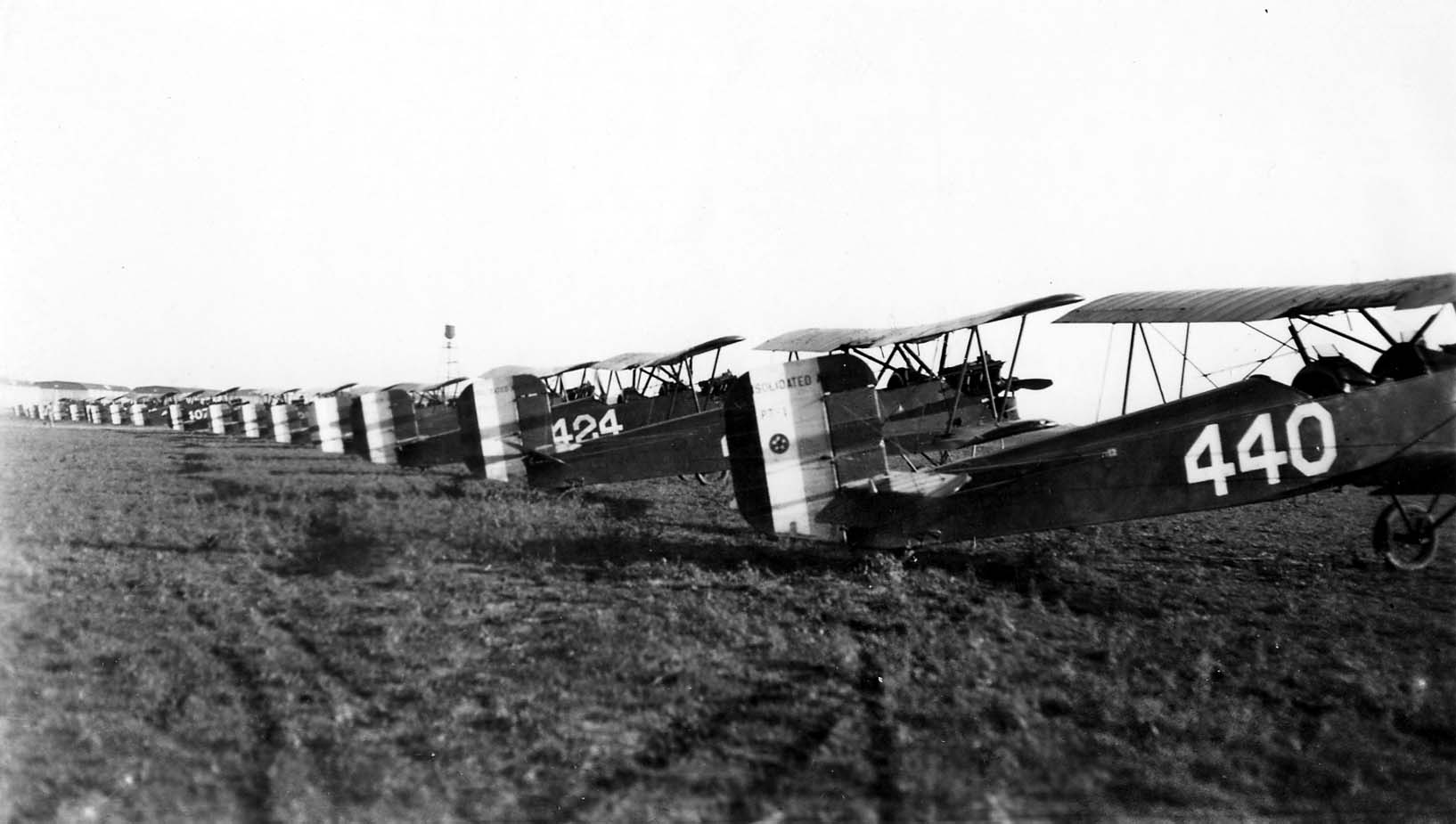 A long, perfectly aligned row of Consolidated PT-1 training biplanes parked on the grassy flight line at Brooks Field.