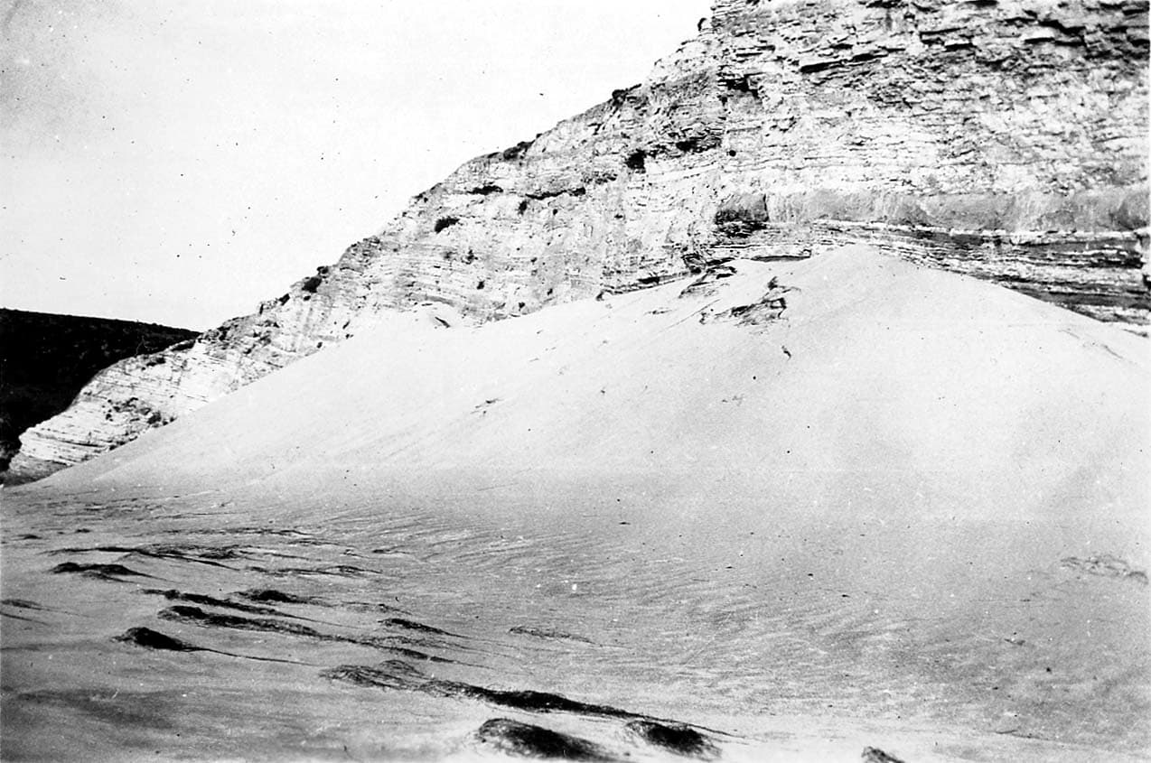A smooth sand dune sweeps up against the base of layered, weathered rock cliffs in an arid desert landscape.