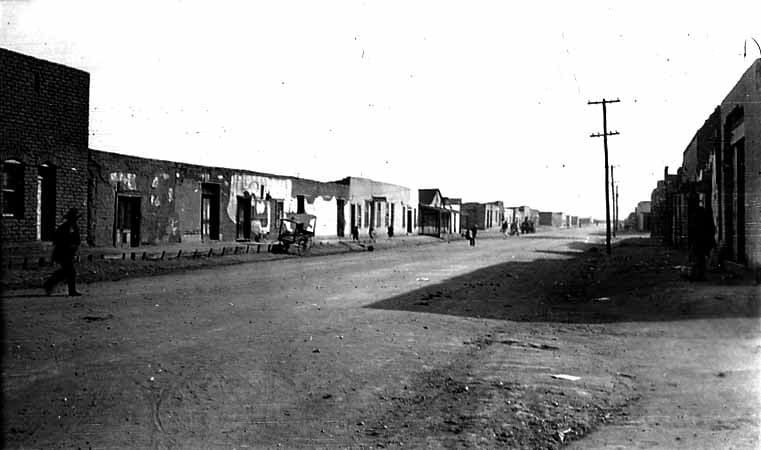 A wide view looking down a wide, unpaved dirt street lined with flat-roofed adobe buildings and wooden porticos in a sun-baked desert town.