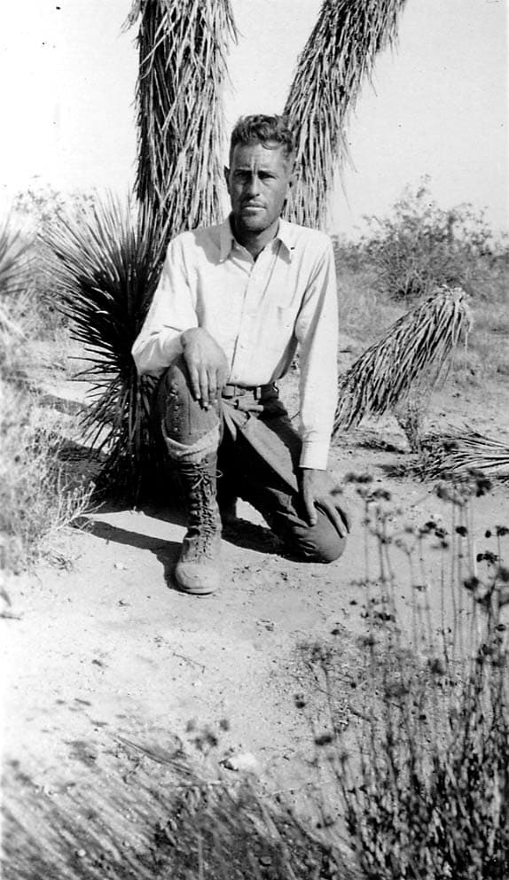 Donald A. Hall Sr. kneeling in the desert sand in front of yucca plants, wearing a light shirt and tall lace-up hiking boots.