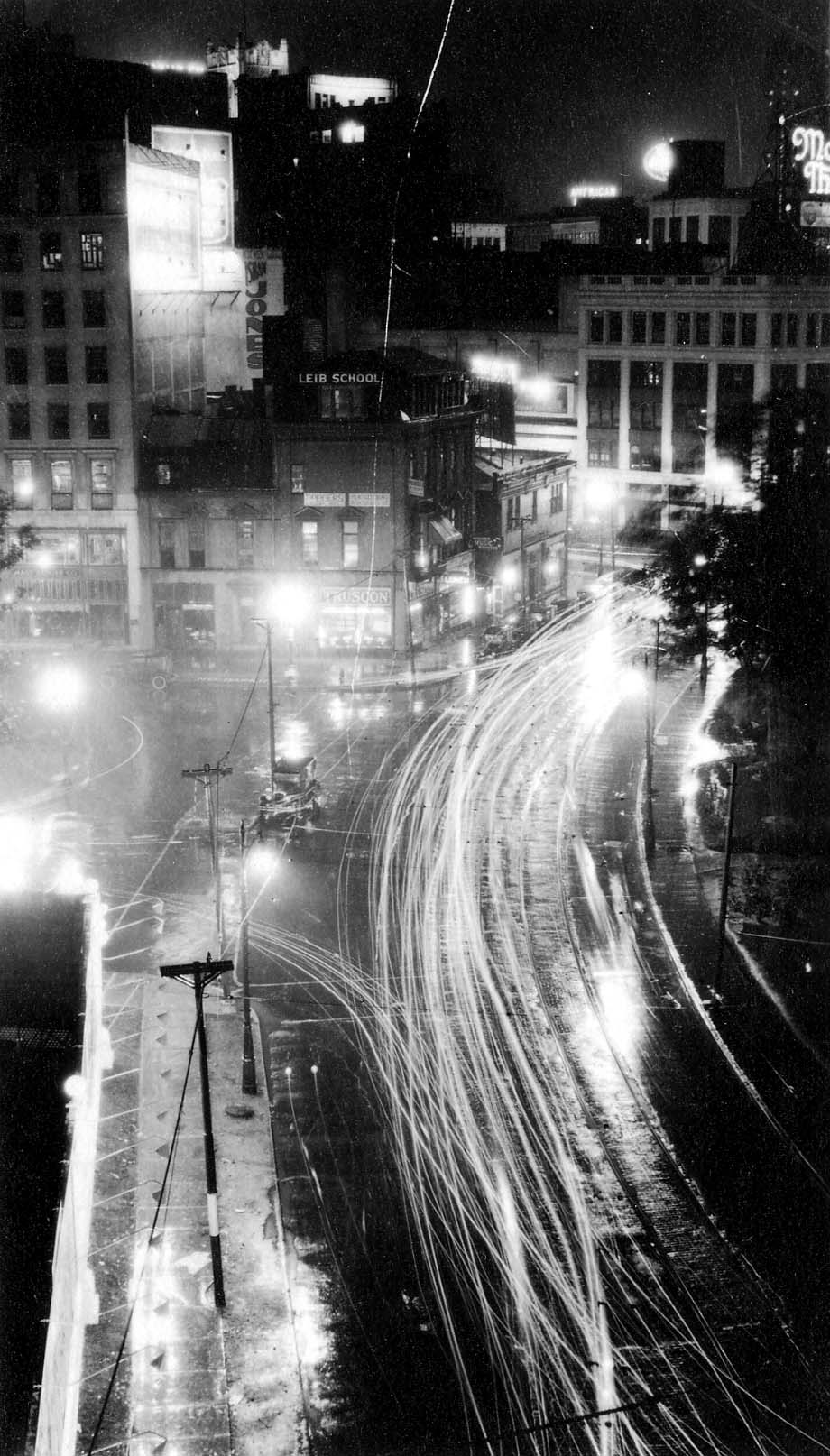 A high-angle nighttime view looking down from an upper window onto rain-slicked Detroit streets, showing sweeping light trails from passing streetcars and automobiles beside the Truscon storefront.