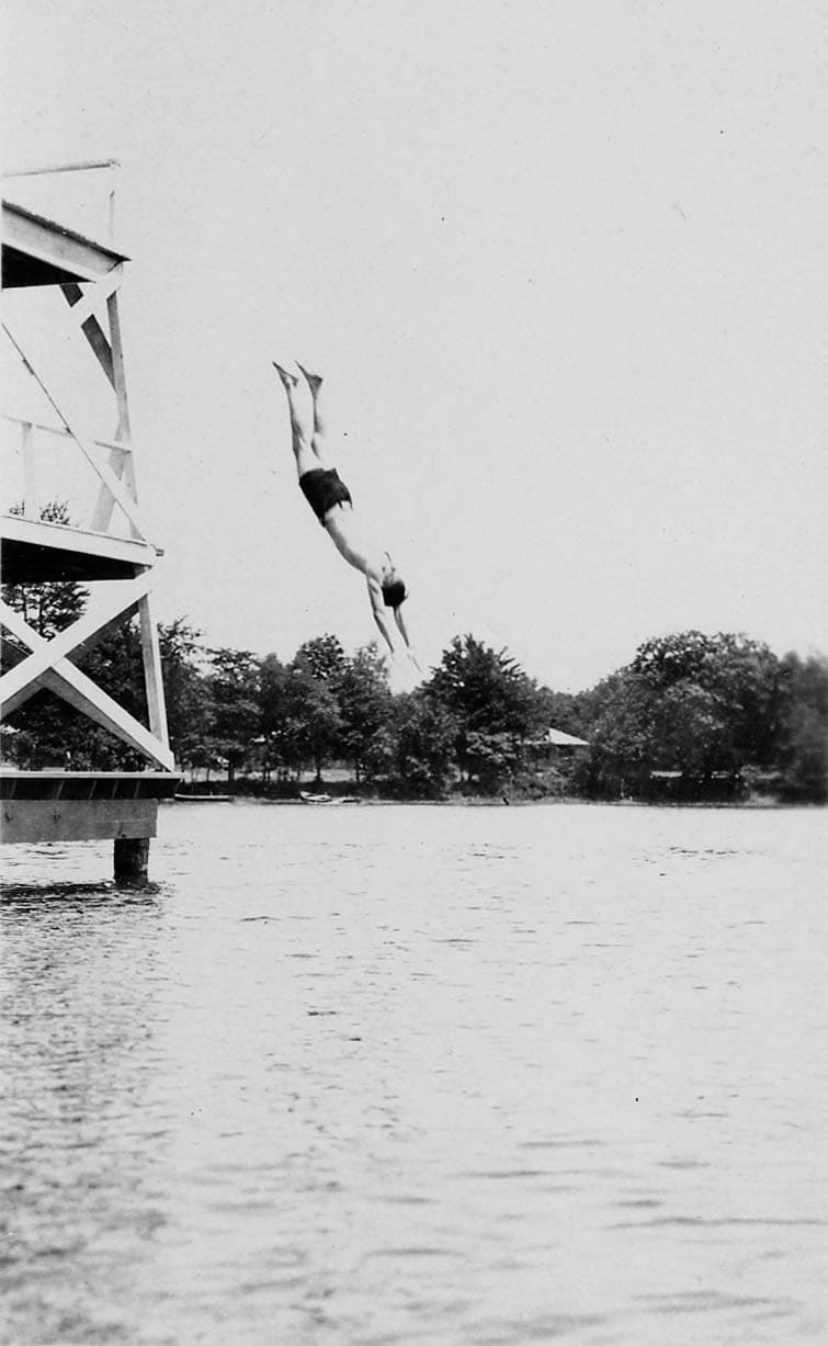 An action shot of a man executing a dive from a high, wooden diving tower into a body of water, with trees visible on the far shore.