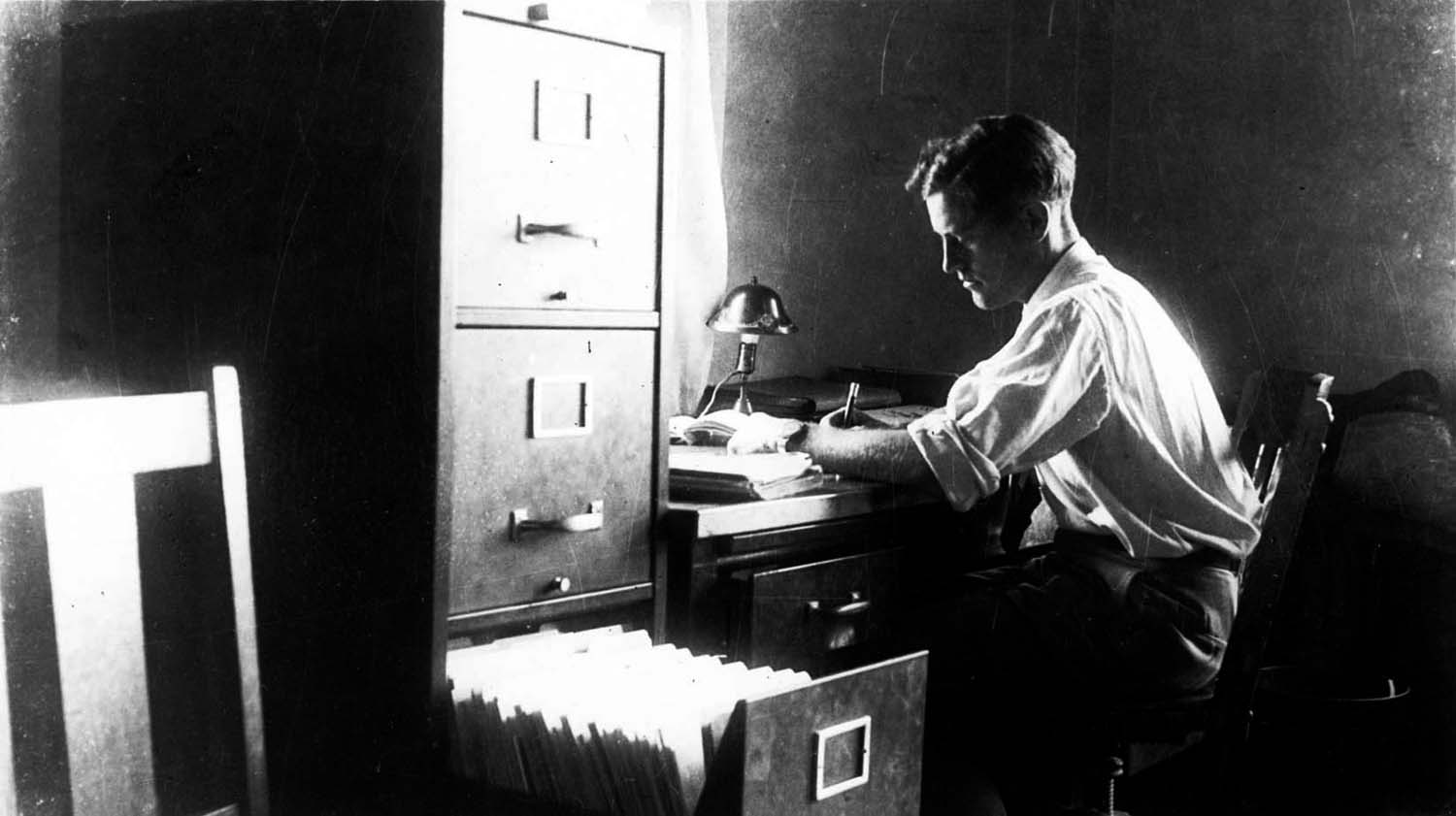 A candid self-portrait of Donald A. Hall Sr. working late at his desk beside a massive metal filing cabinet.