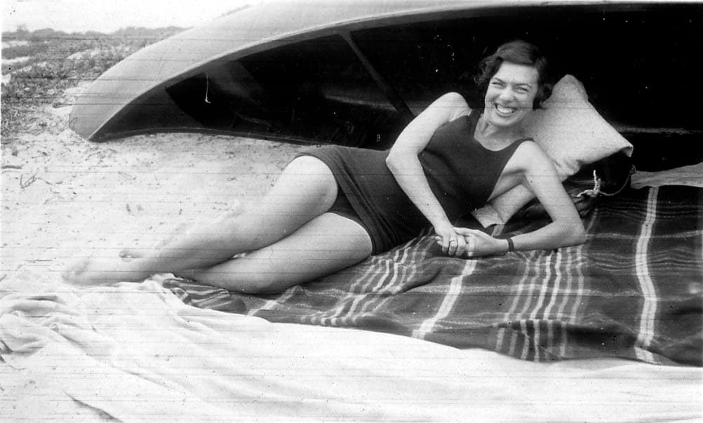 A playful portrait of Elisabeth "Betty" Hall smiling broadly, wearing a dark swimsuit and reclining on a blanket in the sand next to an overturned boat.