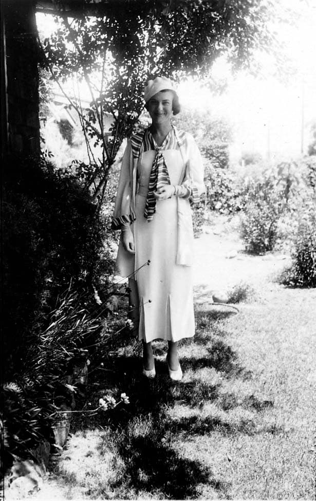 A full-length portrait of Elisabeth "Betty" Hall standing in a sunny garden, wearing a light-colored dress and a dark coat, smiling warmly.
