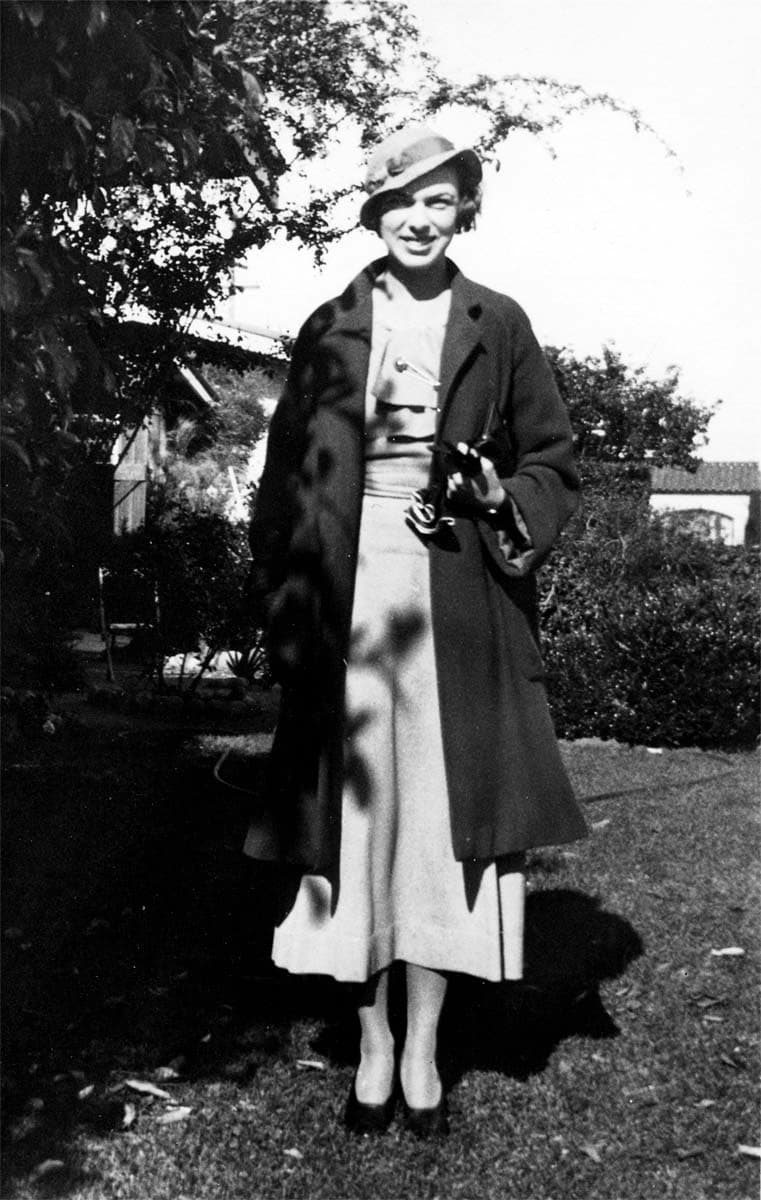 A portrait of Elisabeth "Betty" Hall standing in a sunny garden, wearing a light-colored dress, a dark coat, and a wide-brimmed hat.