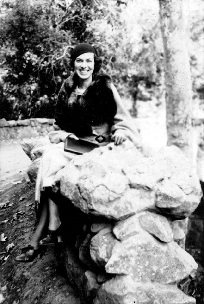 A portrait of Elisabeth "Betty" Hall smiling warmly, wearing a cloche hat and a fur-trimmed coat, sitting on a rustic stone wall.