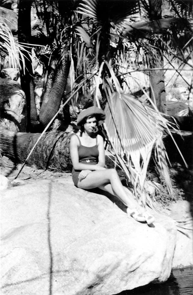 A portrait of Elisabeth "Betty" Hall wearing a wide-brimmed hat and swimsuit, sitting on a large rock surrounded by palm trees and rugged terrain.
