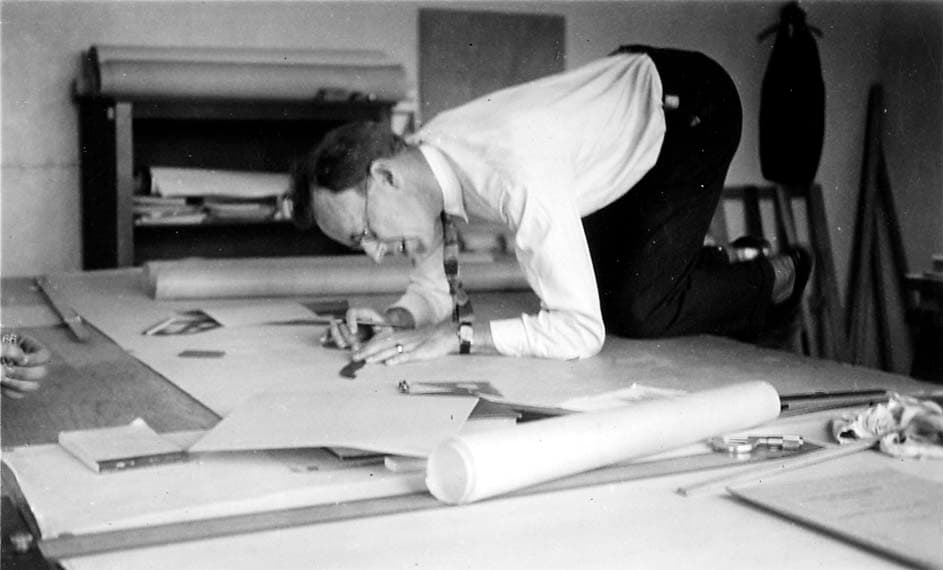 An engineer wearing a shirt and tie kneeling directly on top of a massive architectural blueprint spread across a large drafting table.