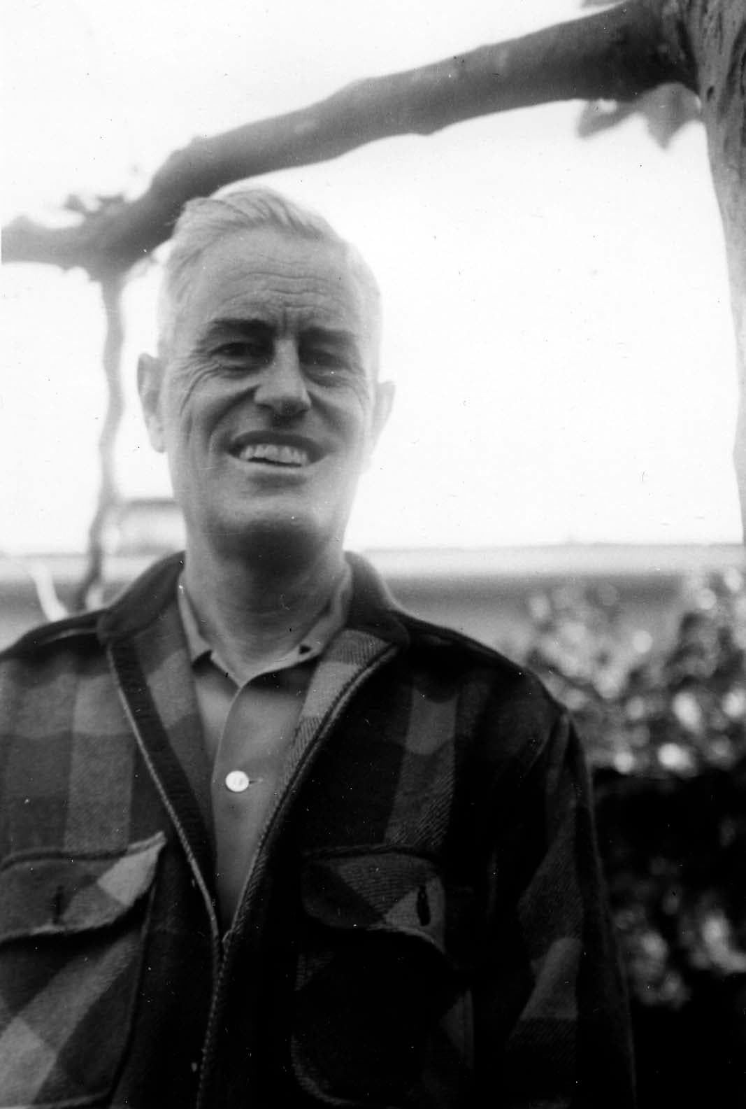 An older gentleman smiling warmly outdoors, wearing a heavy plaid wool jacket over a collared shirt.