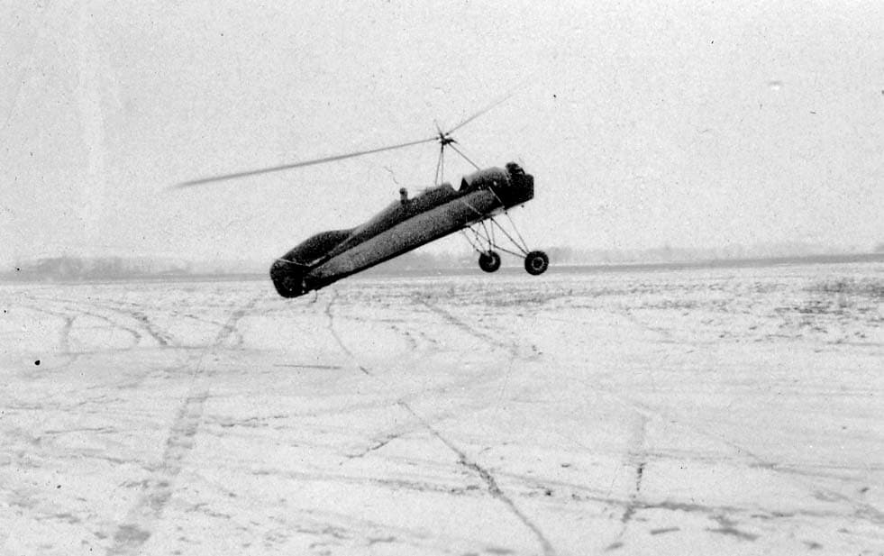 An experimental autogyro flying low over a snow-covered field marked with tire tracks.
