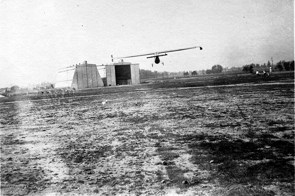 A very lightweight, high-wing experimental aircraft or glider flying low over a grassy airfield, with large hangars in the background.