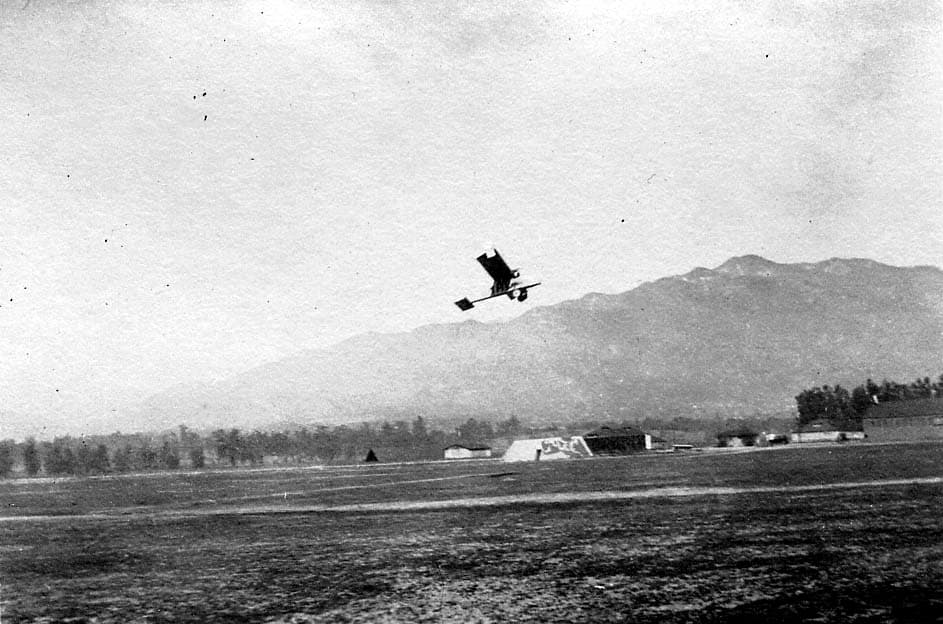 A lightweight, experimental high-wing aircraft or glider flying low over a dirt airfield, with a rugged mountain range in the background.