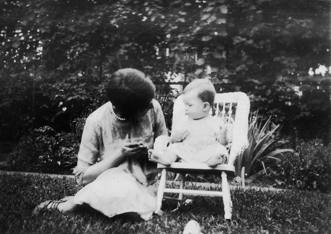 A woman sitting on the grass in a sunny yard, gently holding the hands of a young baby seated in a white wicker highchair.